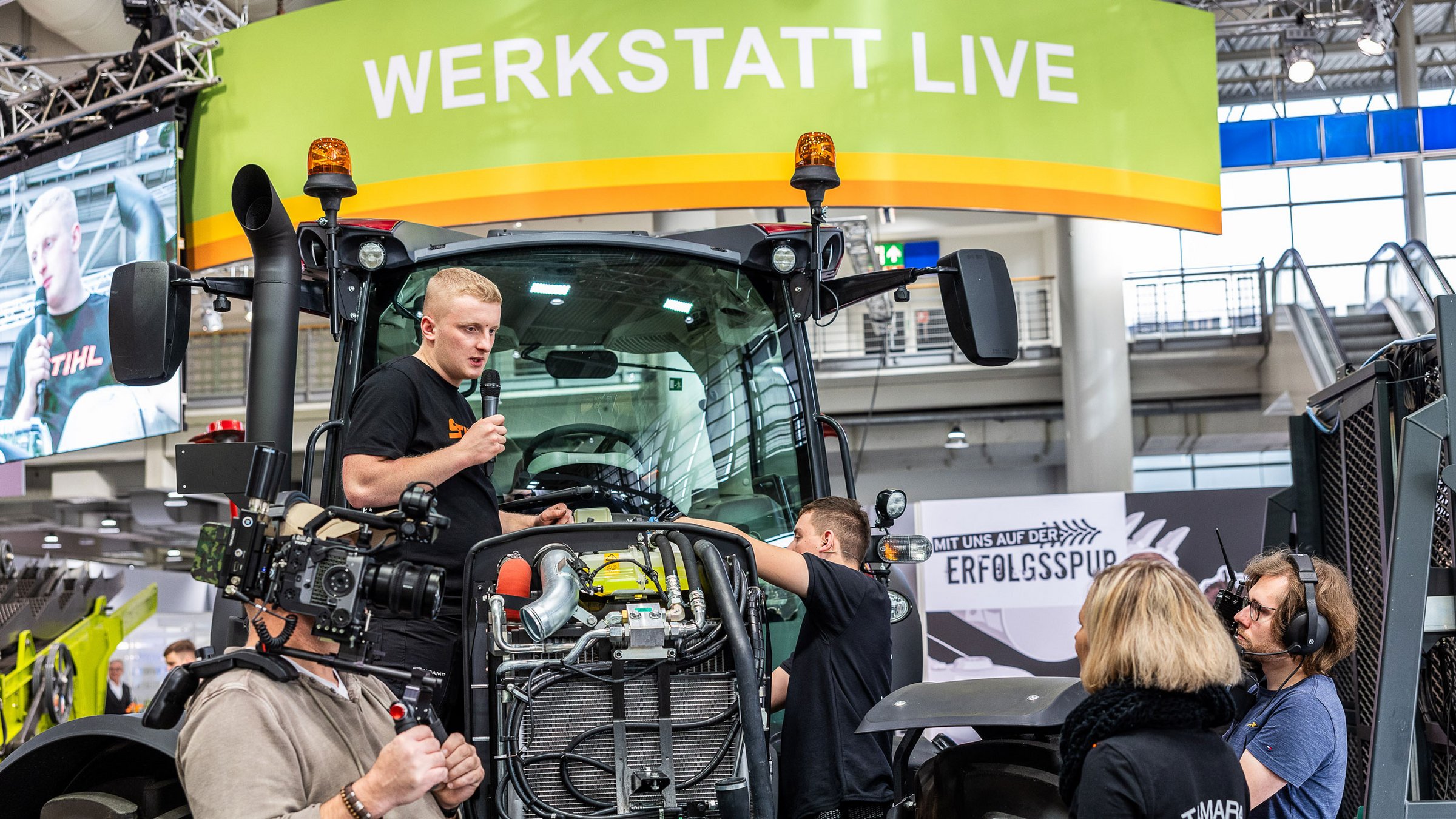 Junger Mann stehend auf Traktor gibt Interview für Filmcrew in einer Messehalle